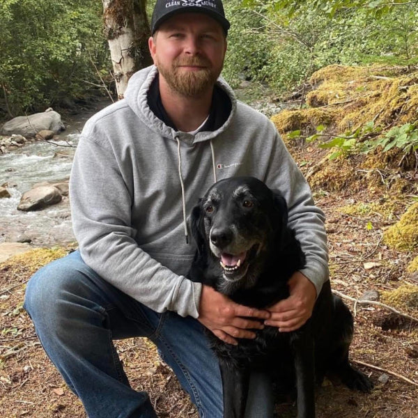 Aaron in a gray hoodie and cap sitting with a black dog outdoors near a stream.